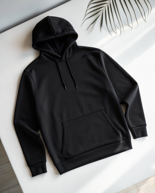 A black pullover hoodie with a front kangaroo pocket and drawstrings, laid flat on a white surface with a palm leaf in the upper right corner.