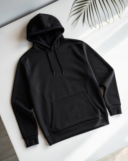 A black pullover hoodie with a front kangaroo pocket and drawstrings, laid flat on a white surface with a palm leaf in the upper right corner.